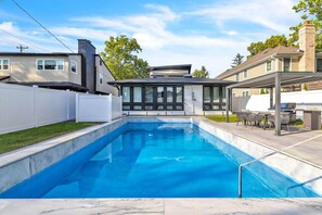 A rectangular swimming pool with a tiled edge, a covered patio area with outdoor furniture, and a white fence surrounding the property. - Elmhurst 4BR Home with Pool | Mid-Term Ready (Elmhurst)