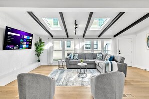 A modern living room with a sectional sofa, a flat-screen TV mounted on the wall, skylights, and a potted plant. - Elmhurst 4BR Home with Pool | Mid-Term Ready (Elmhurst)