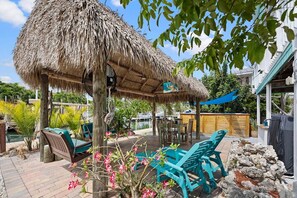 A patio area with a thatched roof, outdoor seating, and a small garden. - Yellowtail Retreat | Canal Front 3BR w/ Dockage, Swim Spa & Gulf Access | Dog-Friendly (Marathon)