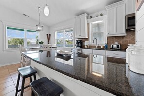 A modern kitchen with white cabinetry, a granite countertop, and a large island with bar stools. - Yellowtail Retreat | Canal Front 3BR w/ Dockage, Swim Spa & Gulf Access | Dog-Friendly (Marathon)