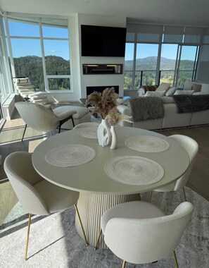 A modern living room with a round dining table, white chairs, a fireplace, and a large window with a mountain view.