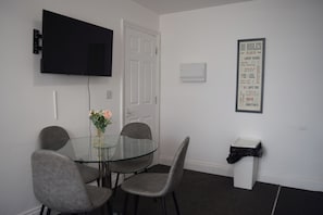 Un comedor con una mesa de vidrio, cuatro sillas grises, un televisor negro montado en la pared y una lista de normas enmarcada en la pared.
