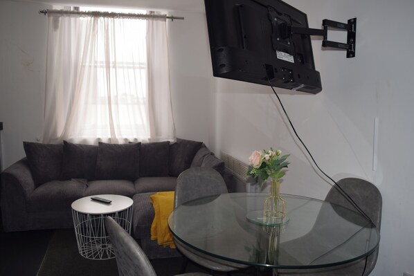 A living room with a sofa, a glass table, a remote control, a flower vase, and a mounted TV.