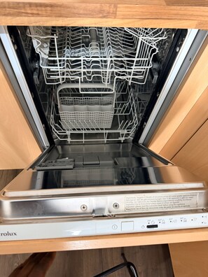 Un lave-vaisselle ouvert, avec ses grilles et ses paniers à l’intérieur, un panneau de commande avec différents réglages, et une porte de meuble en bois.