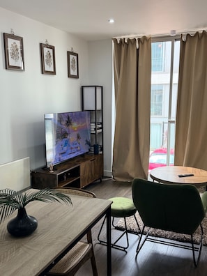 Ein Wohnzimmer mit Fernseher, einer Holzkassette, einem Esstisch mit Stühlen und einem Fenster mit Vorhängen.