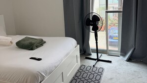 A hotel room with a bed, a standing fan, a window with curtains, and a view of the outside. - Central 1-Bed Apt, Sleeps 2 w/ Underground Parking (Birmingham)