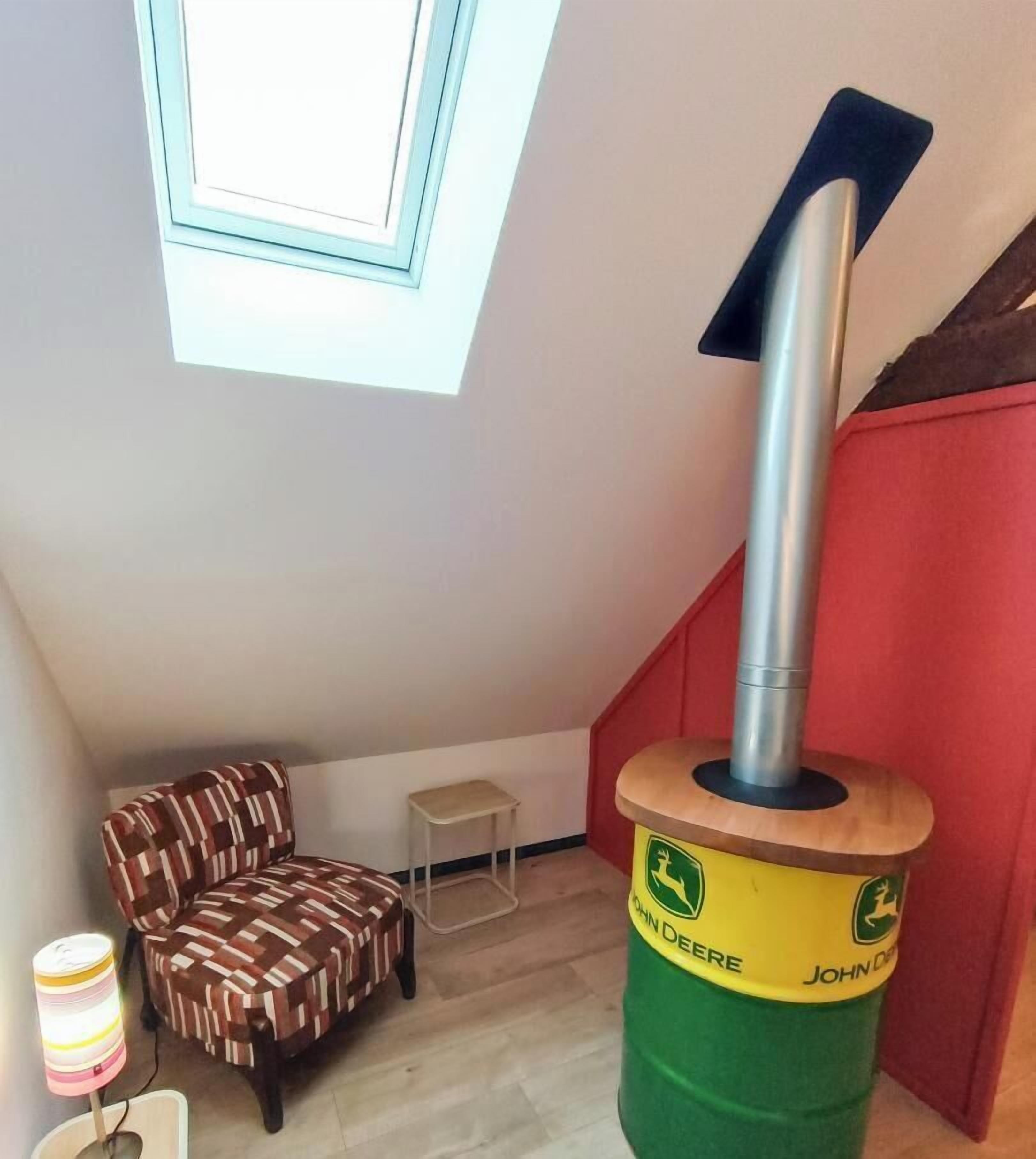 Une petite pièce avec un plafond en pente, une chaise à motifs, une petite table et un poêle à bois John Deere.