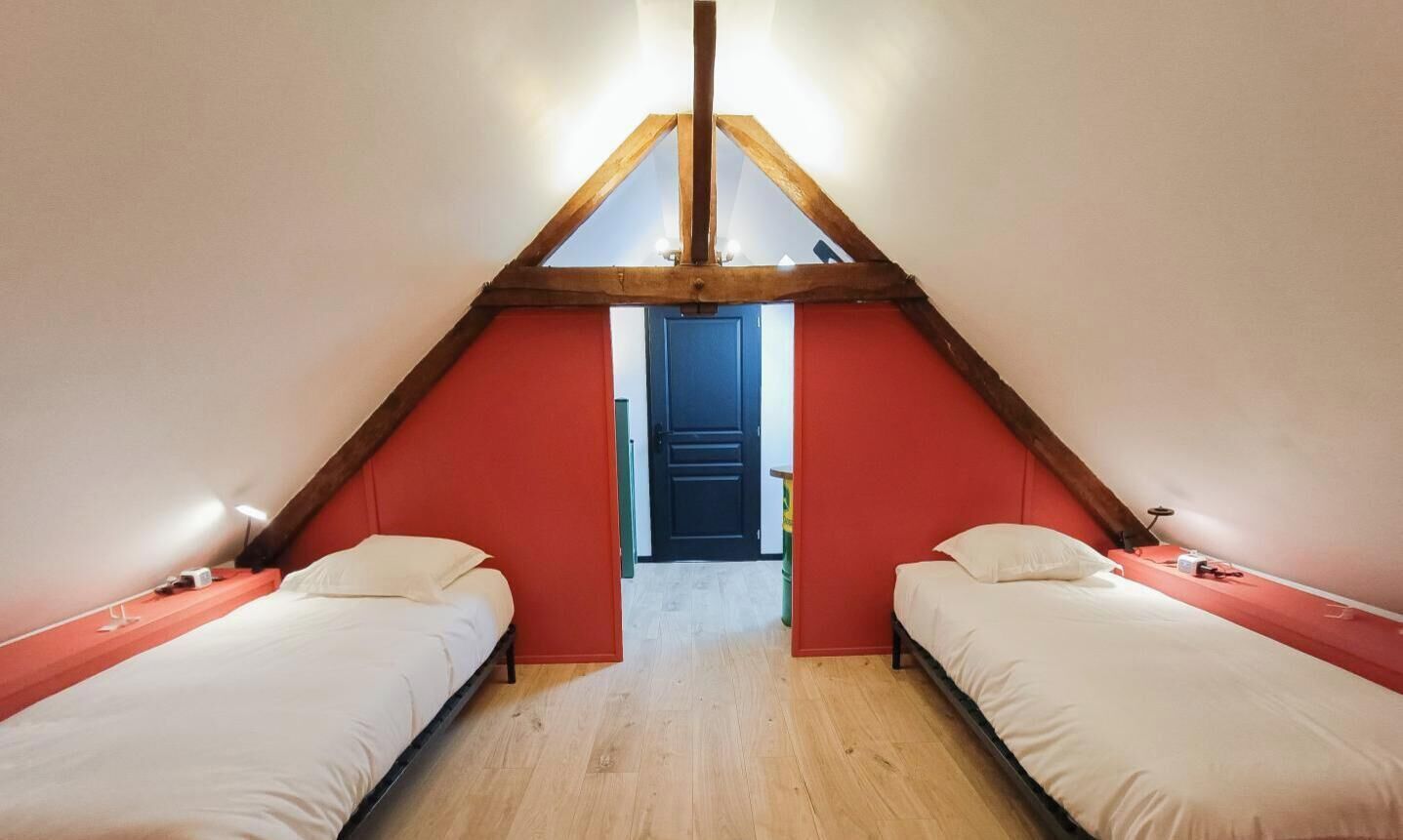 Une chambre mansardée triangulaire, avec deux lits, une poutre en bois au plafond et une porte située au centre.