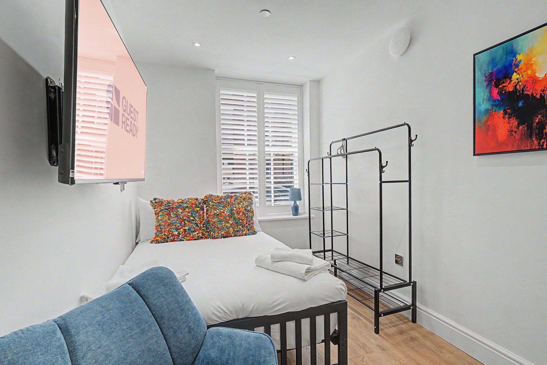 Un dormitorio con una cama, una silla azul, un tendedero de metal, una ventana con persianas y un cuadro colorido en la pared.
