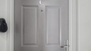 En vit dörr med ett silverfärgat handtag och ett nyckelhål, ett svart föremål på väggen till vänster och en dörrklocka ovanför handtaget.