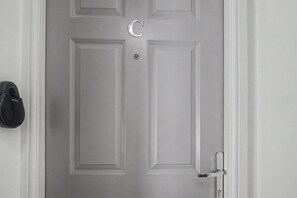 A white door with a silver handle and a keyhole, a black object on the wall to the left, and a doorbell above the handle. - GuestReady - Bright & Modern Studio in Liverpool (Liverpool)