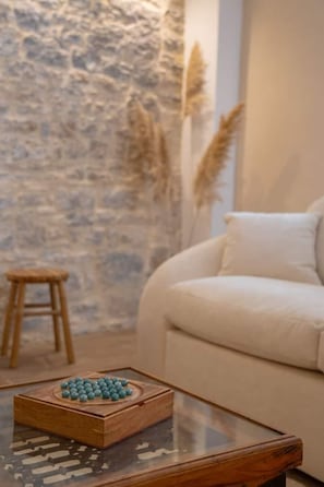 Ein Zimmer mit Steinwand, einem Glastisch mit einer Holzkiste, einem weißen Sofa und einem hölzernen Hocker.