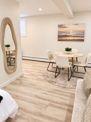 A modern living room with a round dining table, white chairs, a beige sofa, a large mirror, and a beach-themed painting on the wall.