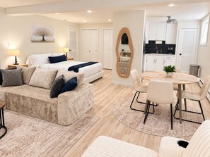 A modern bedroom with a large bed, a sitting area with a sofa and chairs, a dining table with chairs, and a kitchen area with cabinets and a countertop.