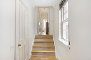 A hallway with white walls, a wooden staircase, and a window on the right. - GuestReady - Charming Flat in Fulham, London (London)