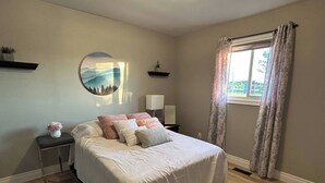 Un dormitorio con cama, mesitas de noche, lámpara de pared, ventana con cortinas y una obra de arte en la pared.