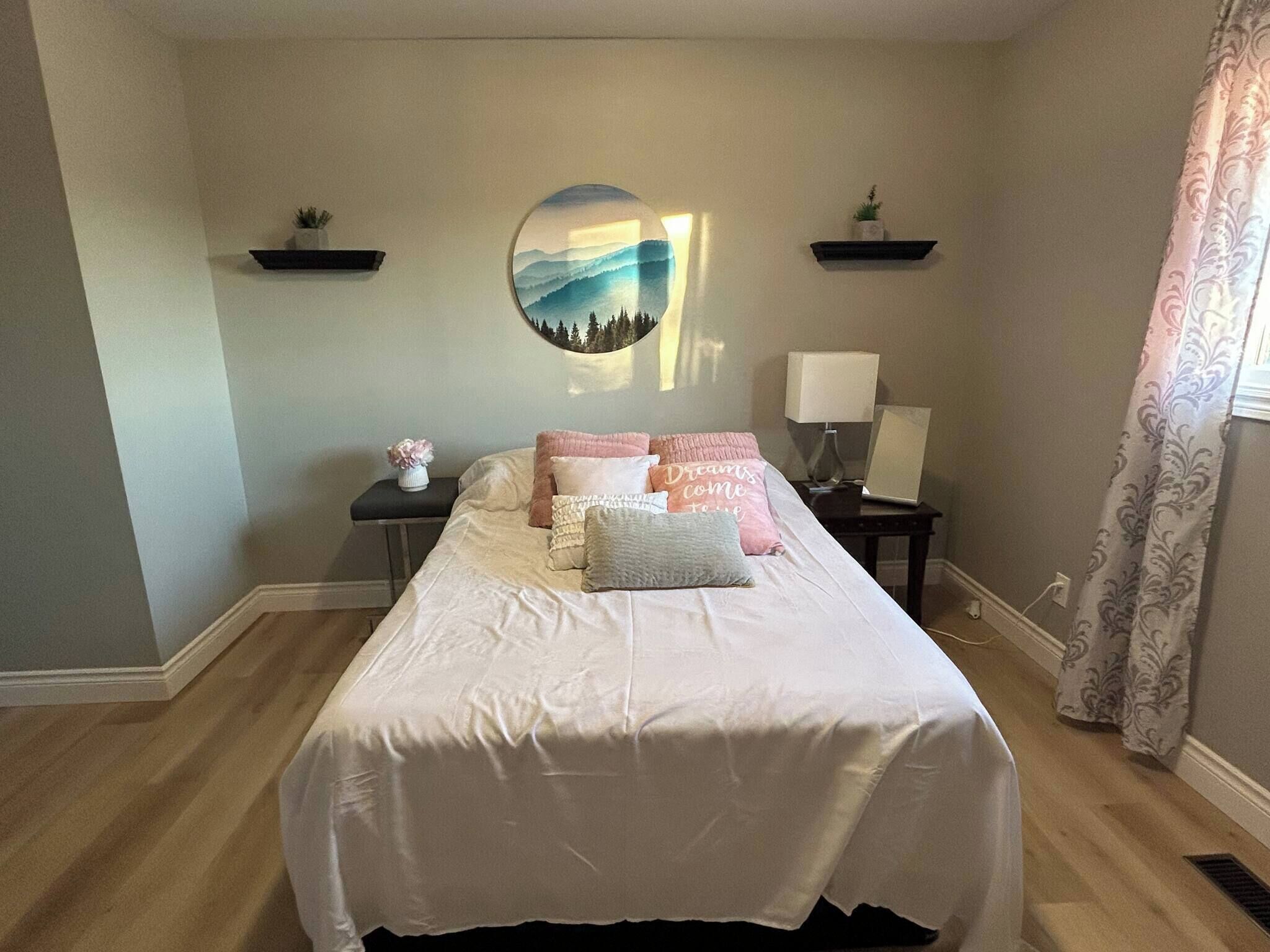 A bedroom with a bed, pillows, a nightstand with a lamp, a wall art piece, and a window with curtains.