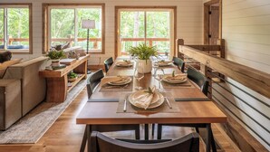 A dining area with a long wooden table set for six, flanked by chairs, with a view of a forest through large windows. - Sauna, Cold Plunge, Hot Tub & Theater - Ellie Blue (Ellijay)