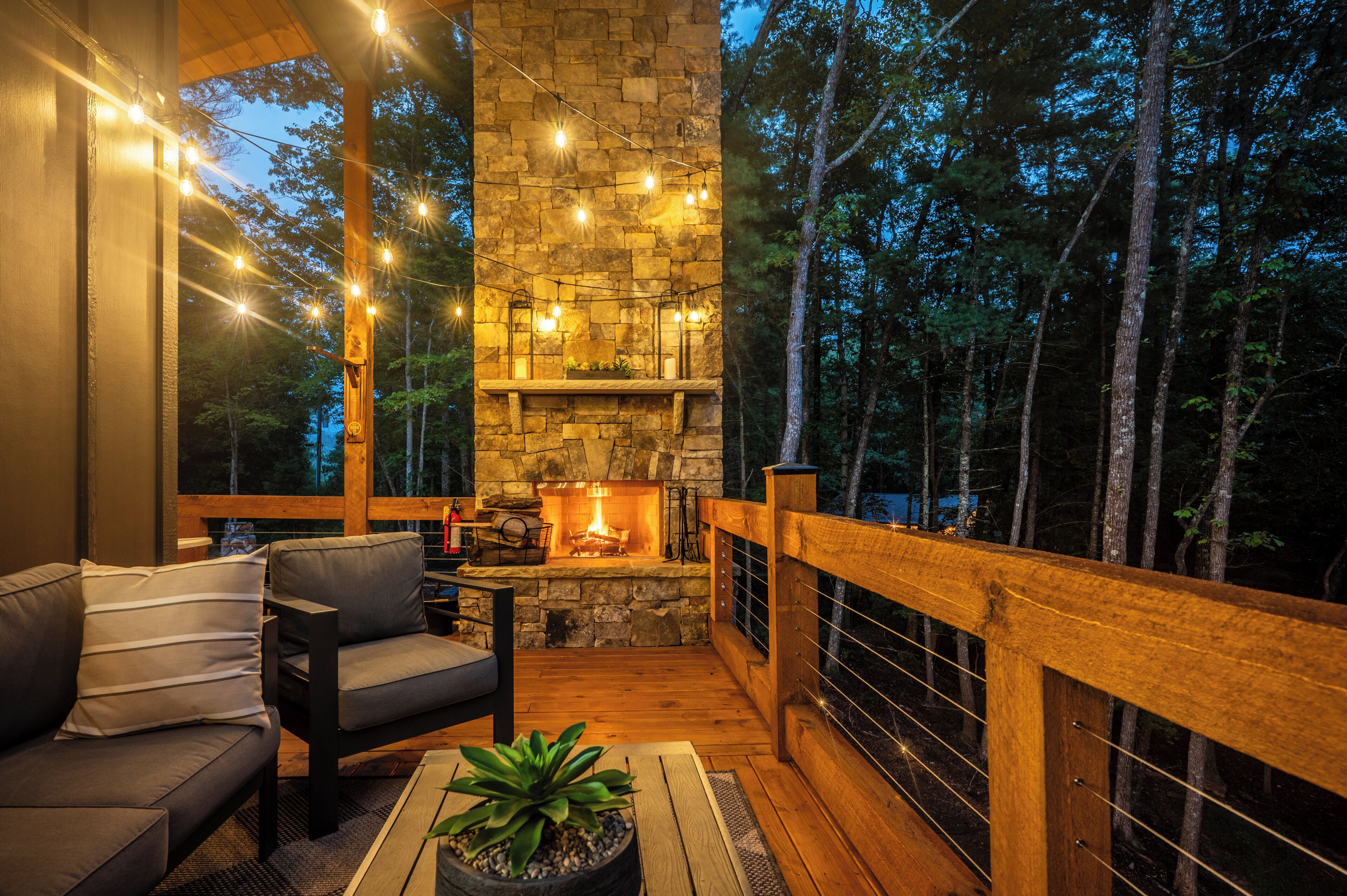 A cozy outdoor seating area with a stone fireplace, wooden deck, and a view of the forest.