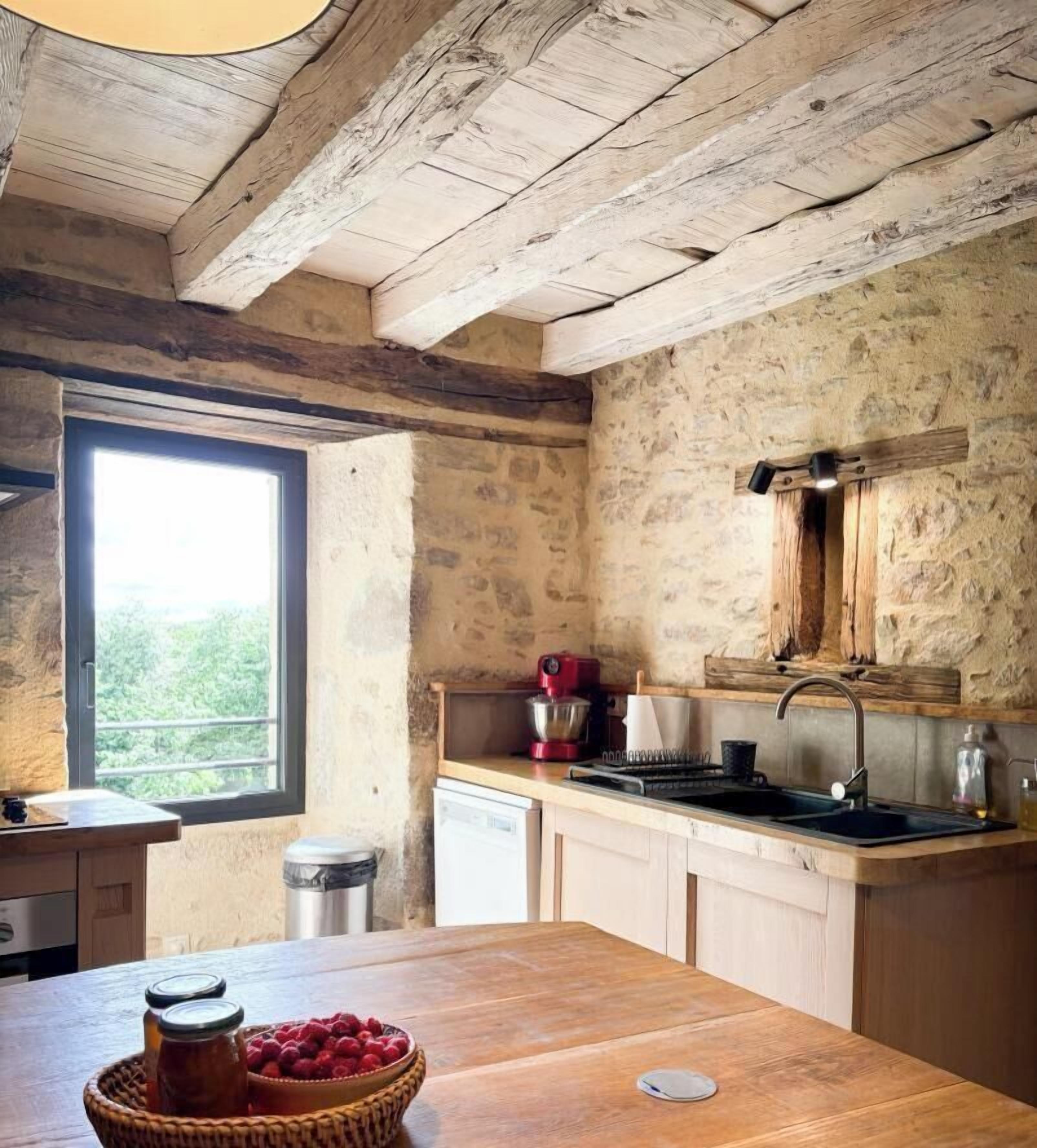 A rustic kitchen with a wooden ceiling, stone walls, a window, and a wooden table with jars and a bowl.