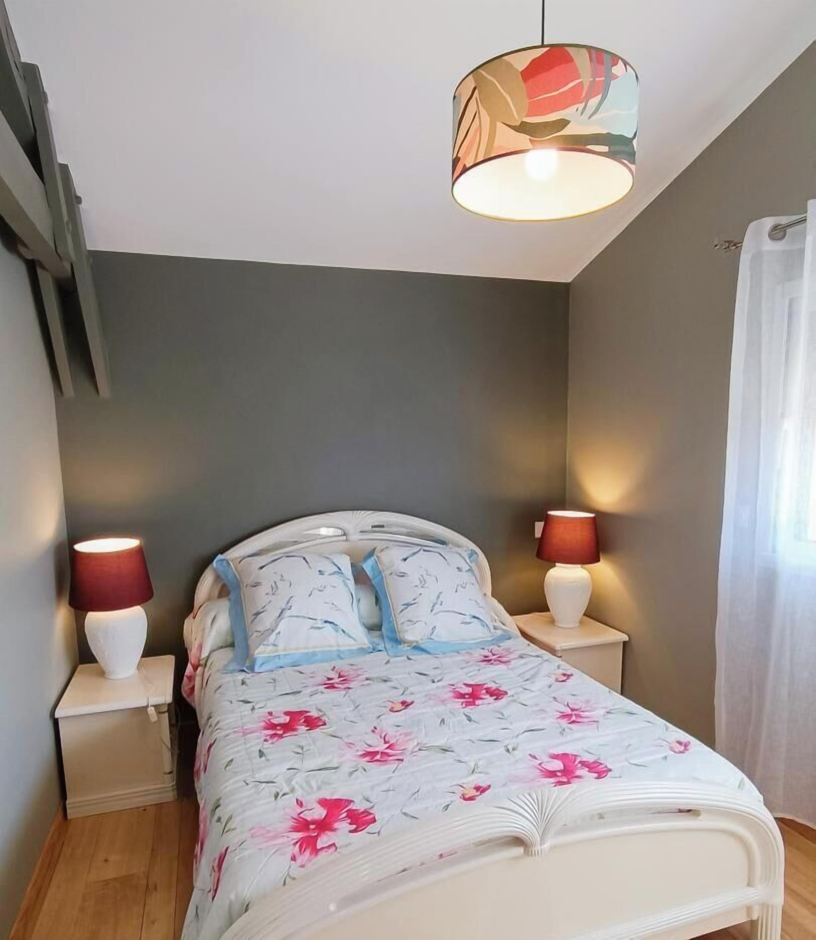 A bedroom with a bed, two bedside lamps, a hanging light fixture, and a window with curtains.