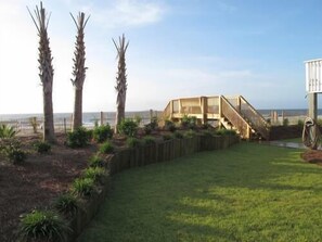 A wooden deck with a staircase leading to a building, surrounded by a grassy area and palm trees. - Alligator Tale - Oceanfront in Garden City Beach (Garden City)