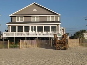 Een huis met twee verdiepingen, een balkon, een houten omheining en een zandstrand op de voorgrond.