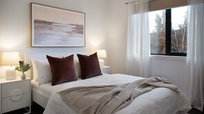 Ein ordentlich bezogenes Bett mit zwei Kissen, ein Nachttisch mit Lampe, ein Fenster mit Vorhängen und ein Bild an der Wand.