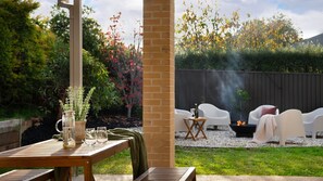A patio with a wooden table, a vase with plants, and a fire pit area with white chairs.