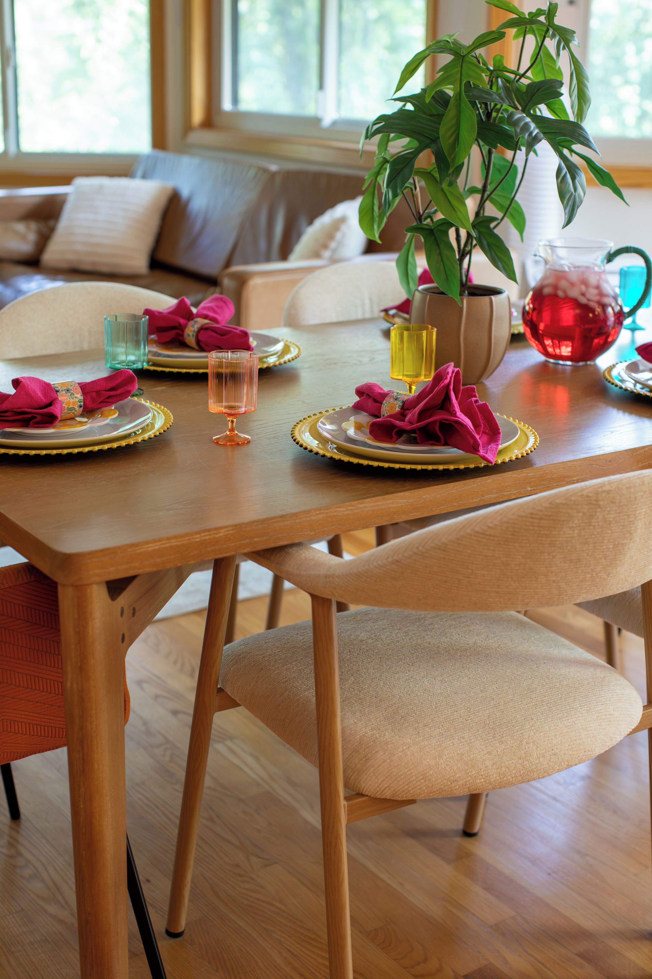 A dining table set with plates, glasses, and napkins, with a plant and a pitcher in the background.