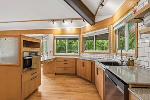 A modern kitchen with wooden cabinets, a stainless steel oven, a dishwasher, and large windows offering a view of greenery.