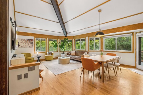 A spacious living area with a dining table, sofas, and large windows offering a view of greenery.