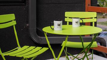 A small outdoor seating area with a table and chairs, a window with blinds, and a black textured wall.