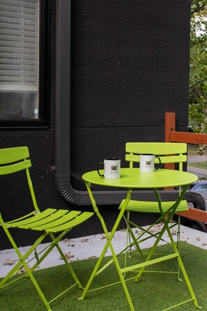 Un pequeño espacio al aire libre con una mesa y sillas, una ventana con persianas y una pared texturizada de color negro.