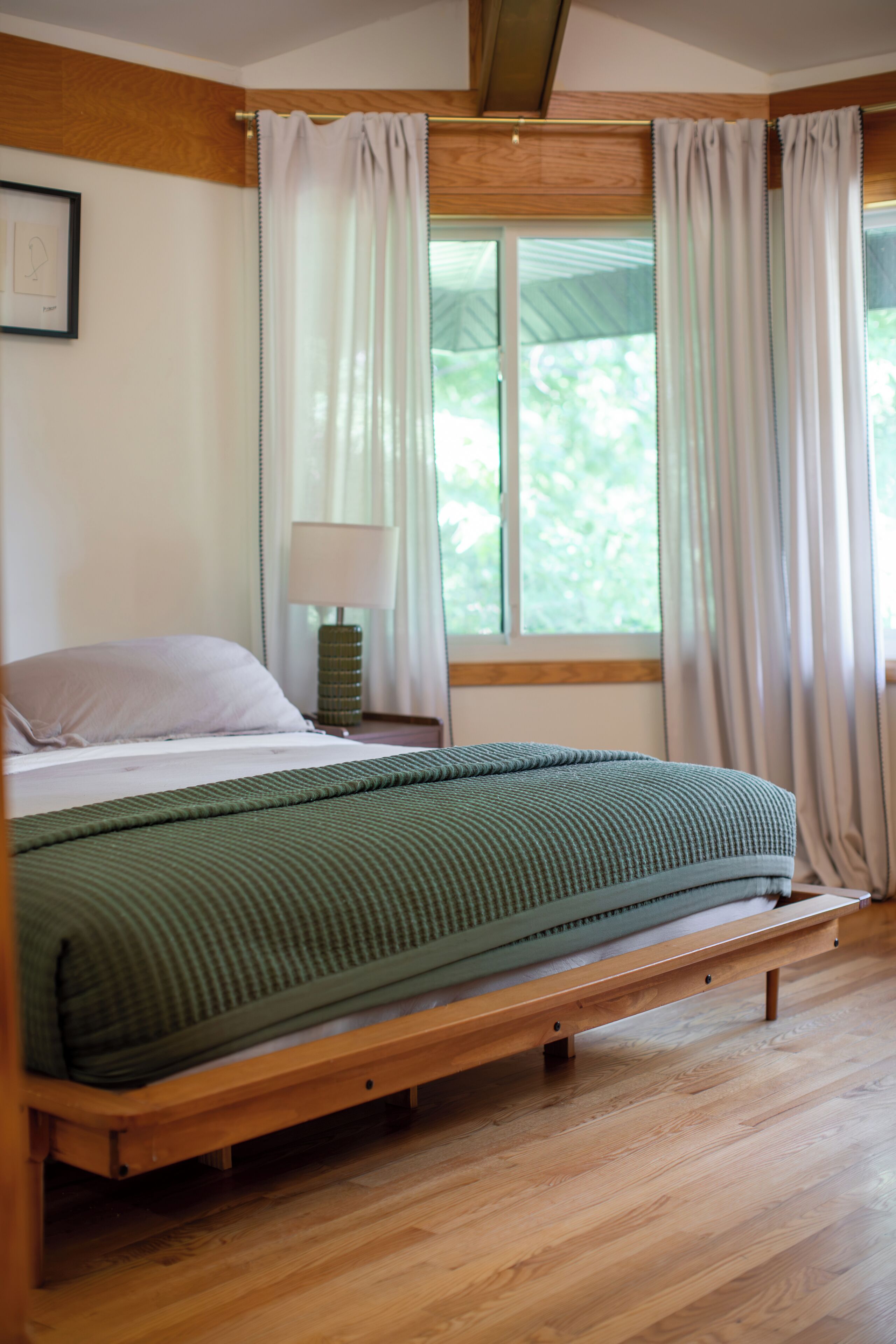 A bedroom with a wooden bed frame, a green bedspread, a bedside lamp, and a window with curtains.