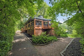 A wooden cabin with a covered porch, surrounded by dense greenery and a gravel path.