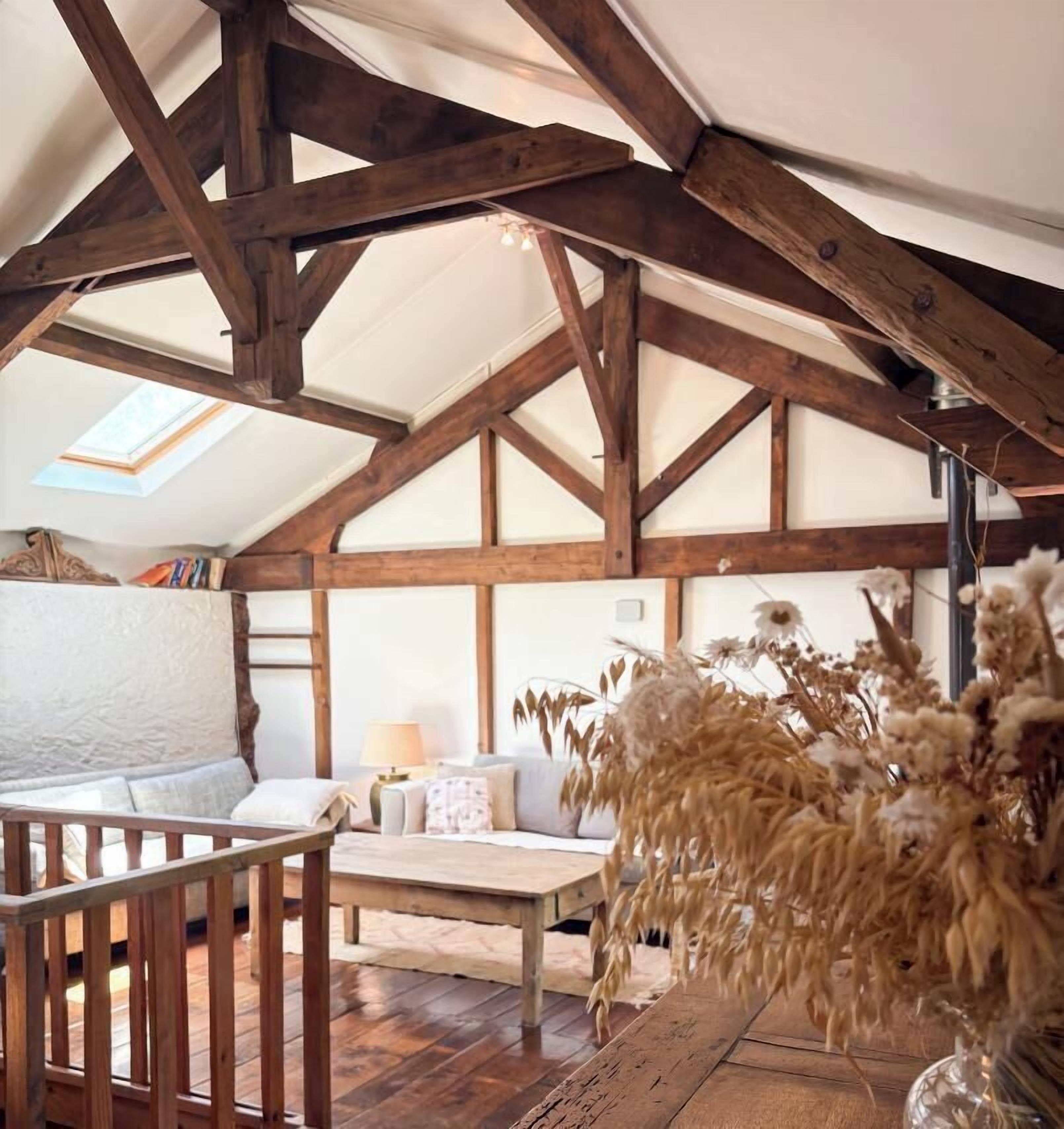 A loft-style room with exposed wooden beams, a skylight, a sofa, a table, and a vase with dried flowers.