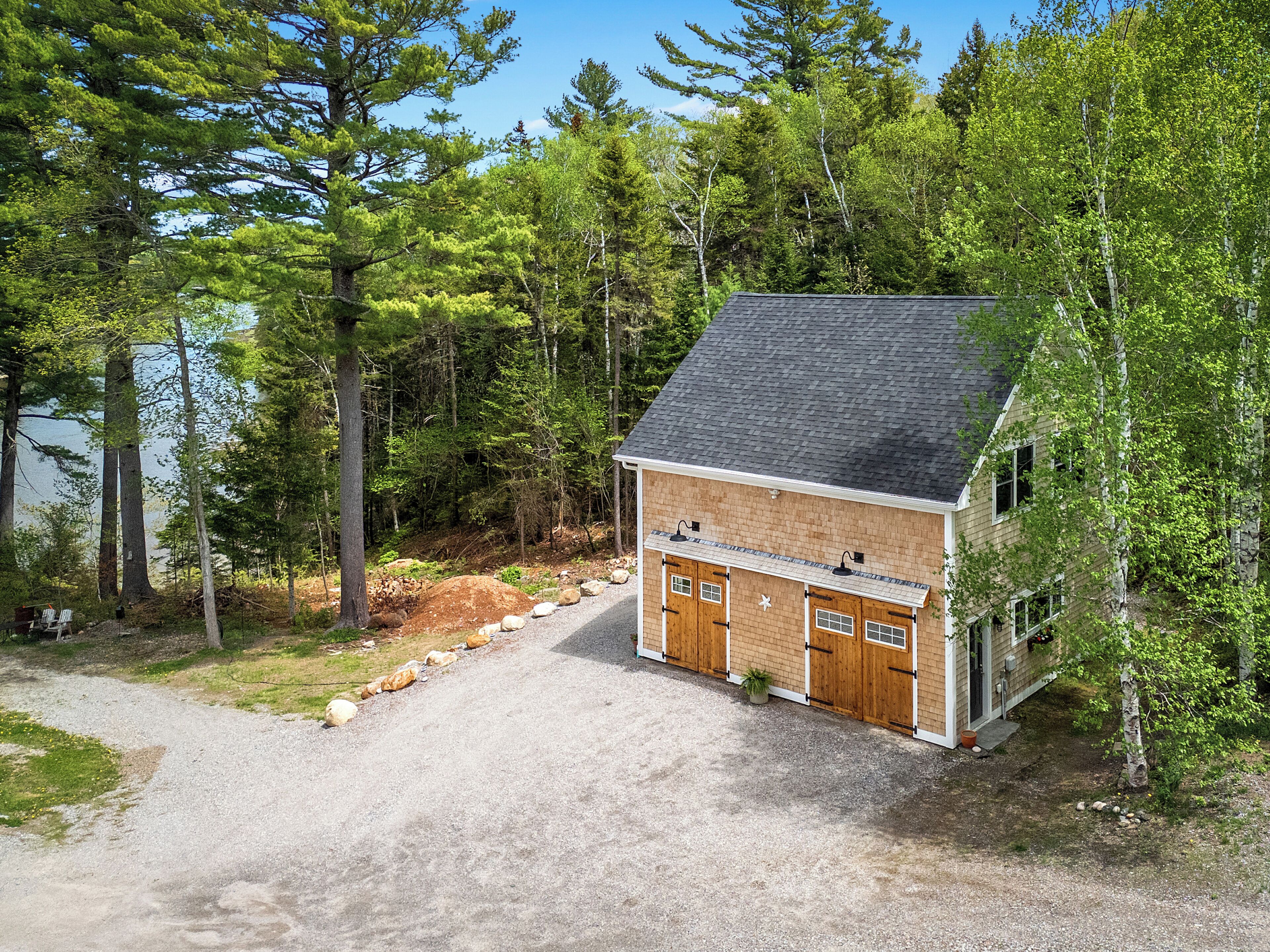 A two-story cabin with a gravel driveway, surrounded by dense forest and a body of water.