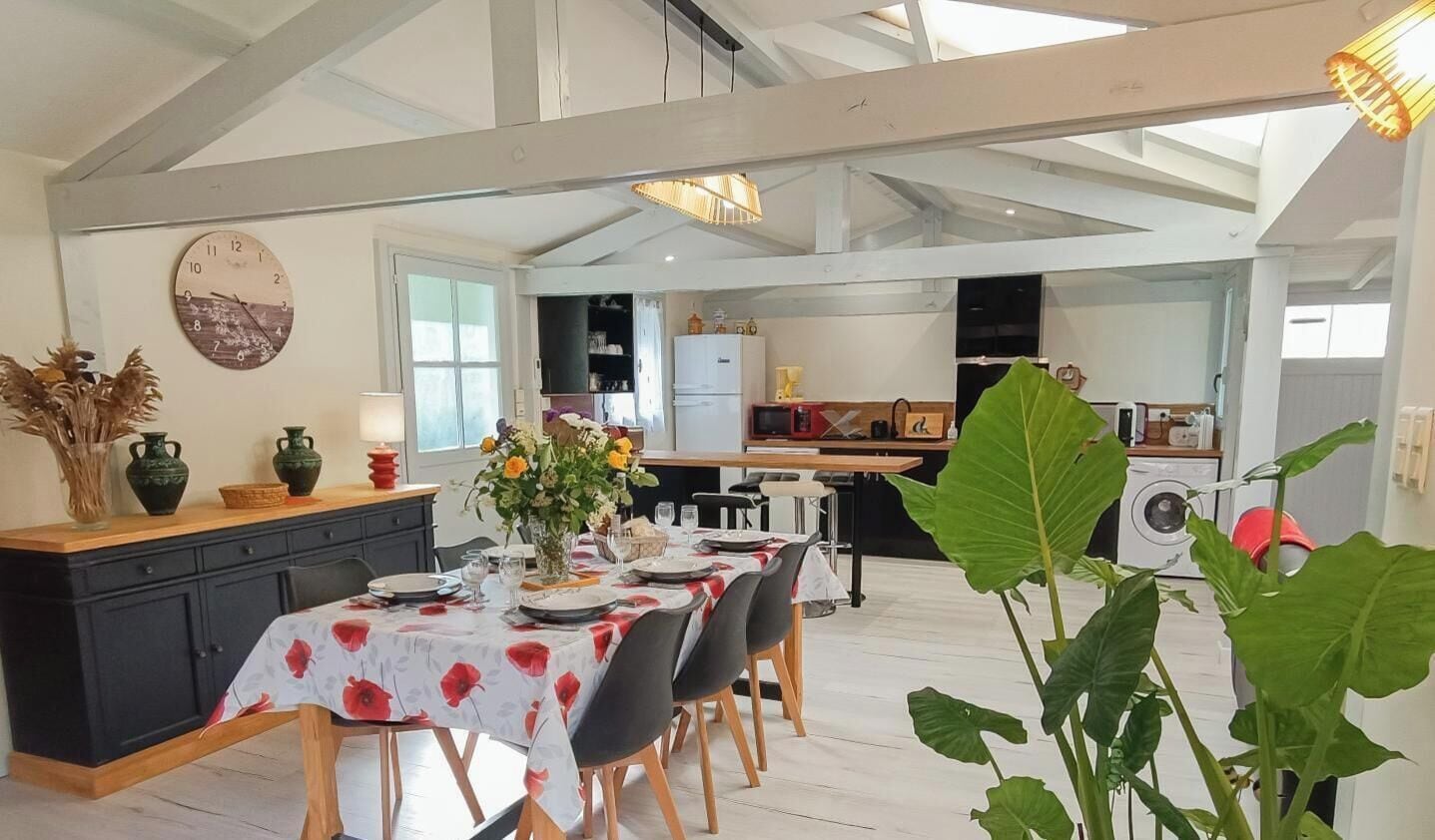 Un coin repas avec une table dressée pour quatre personnes, une cuisine en arrière-plan et une grande plante au premier plan.