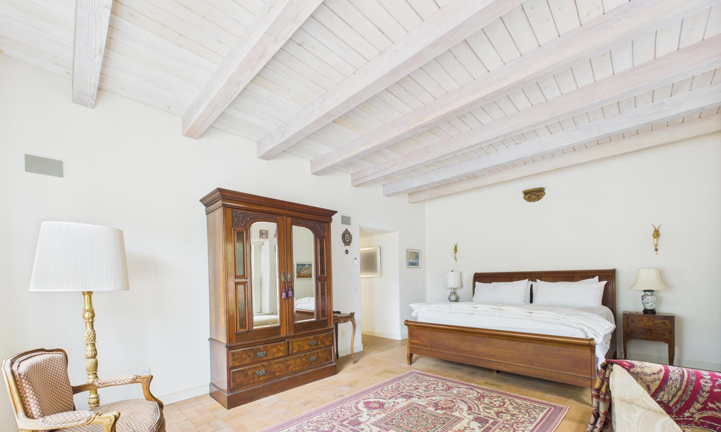 Un dormitorio con cama de madera, un espejo grande, cómoda de madera, una silla y una alfombra estampada.