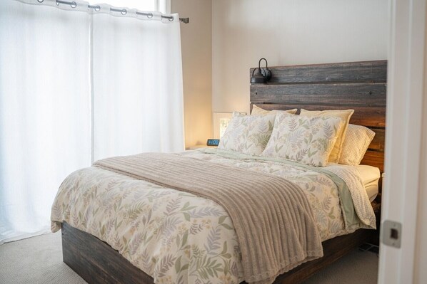 A bedroom with a wooden headboard, a bed with floral bedding, a nightstand with a lamp, and a window with curtains. - Lakefront Mille Lacs Home w/ Dock & Stunning Views (Aitkin)