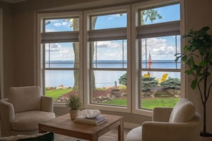 A room with a large window overlooking a lake, a wooden table with books, two armchairs, and a potted plant.
