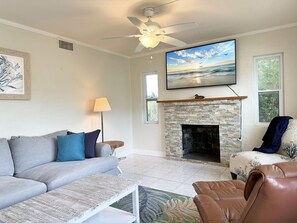 Ein Wohnzimmer mit einer Couch, einem Couchtisch, einem Ledersessel, einem Kamin, einem Deckenventilator und einem Fernseher, der über dem Kamin angebracht ist.