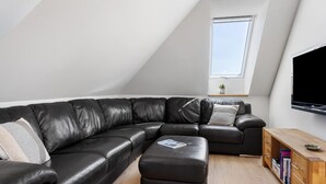 Moderni olohuone, jossa on musta nahkainen kulmasohva, puinen sivupöytä ja seinälle asennettu litteänäyttöinen televisio.