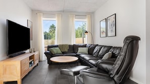 A modern living room with a leather sofa, a flat-screen TV, a wooden coffee table, and a view of the outdoors through large windows. - VillaVilla 981 - Hostrup Strand, Limfjorden (Spøttrup)