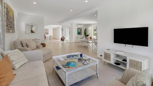Un salon moderne avec une table basse en verre, des canapés blancs, un téléviseur à écran plat et un coin repas à l’arrière-plan.