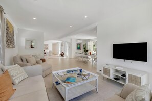 A modern living room with a glass coffee table, white sofas, a flat-screen TV, and a dining area in the background. - Villa Roi Soleil Private pool - St-Tropez, Saint-Tropez, France (Saint-Tropez)