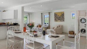 Une cuisine moderne avec des meubles blancs, un coin repas avec une table blanche dressée pour six personnes, et un salon avec un canapé et un tableau.