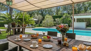 Un espace extérieur aménagé pour les repas, avec vue sur la piscine, comprenant des tables et des chaises en bois, ainsi qu’une terrasse couverte.