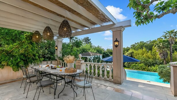Une terrasse couverte avec une table et des chaises, une piscine et une végétation luxuriante.